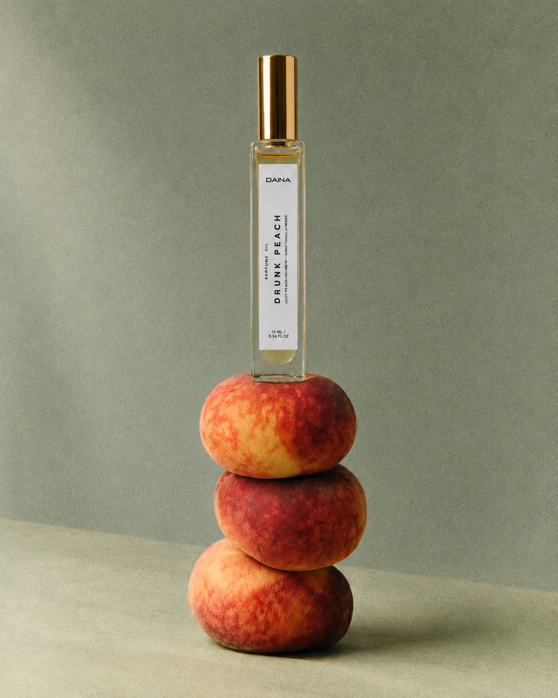 DAINA Drunk Peach perfume bottle with gold cap standing on a stack of three whole peaches against a sage-green background