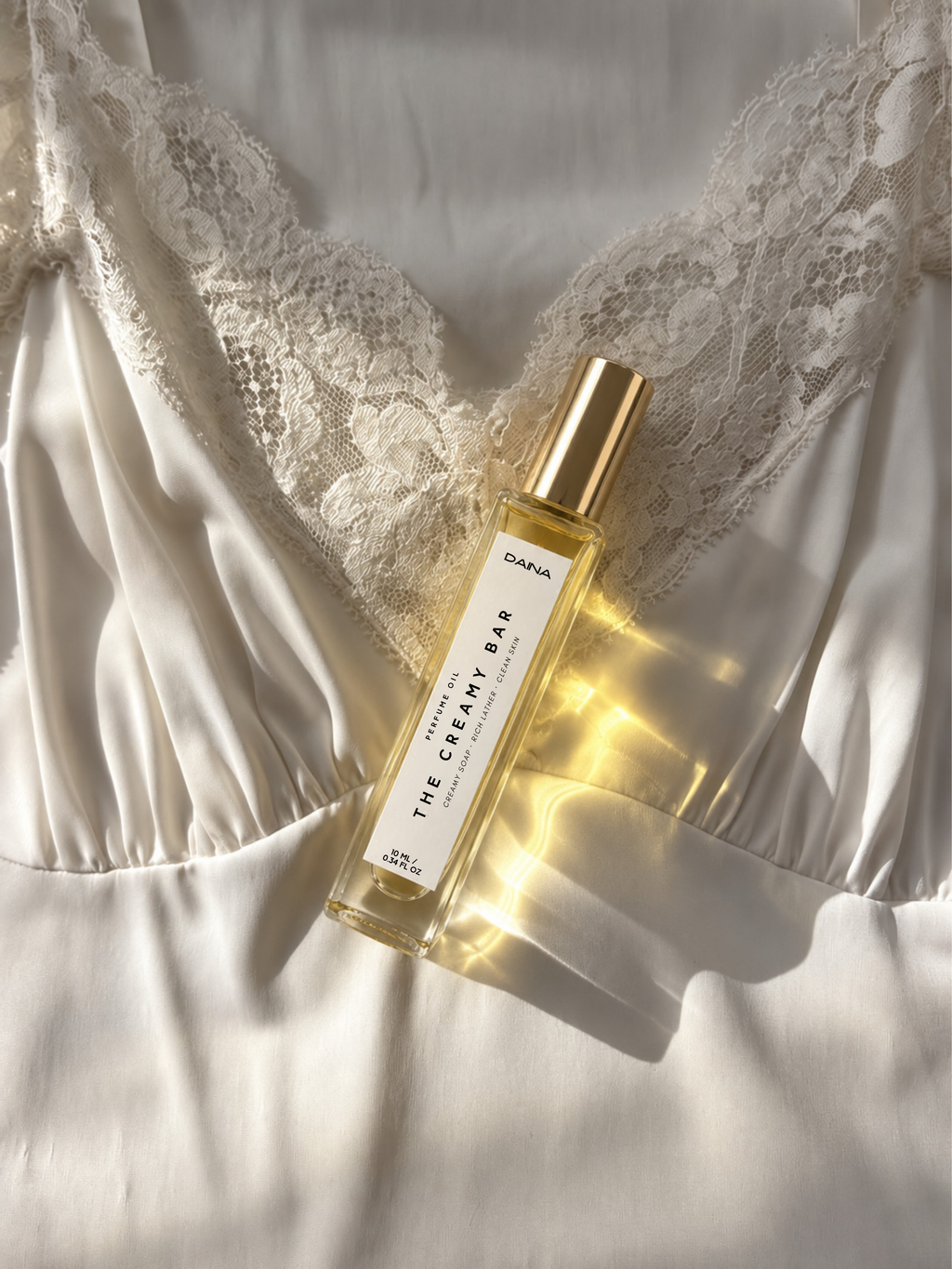 DAINA The Creamy Bar perfume oil, a creamy white soap-inspired fragrance in a 10 ml roller bottle resting on ivory satin and lace in warm sunlight.